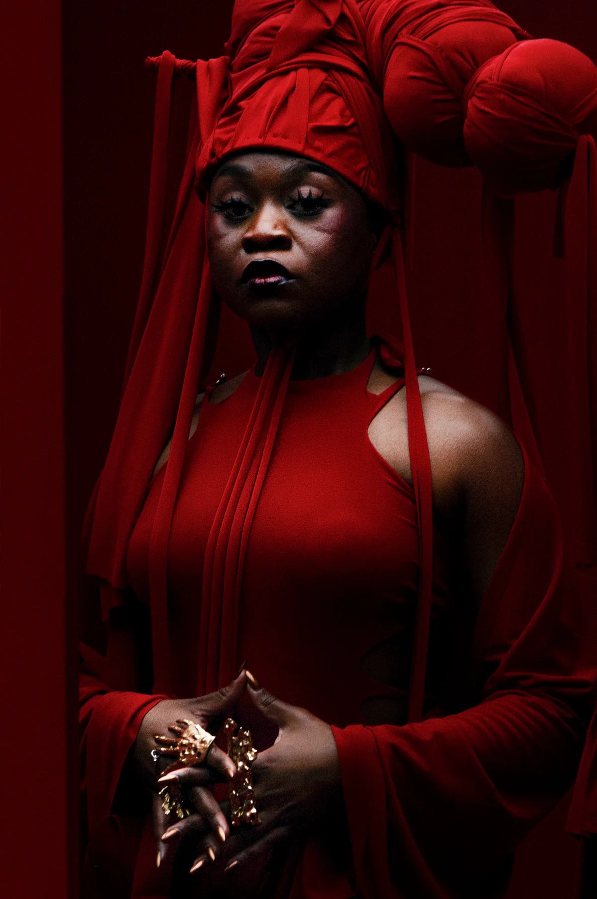 Sampa the Great in a sculptural red headwrap and red garment against a deep red backdrop.
